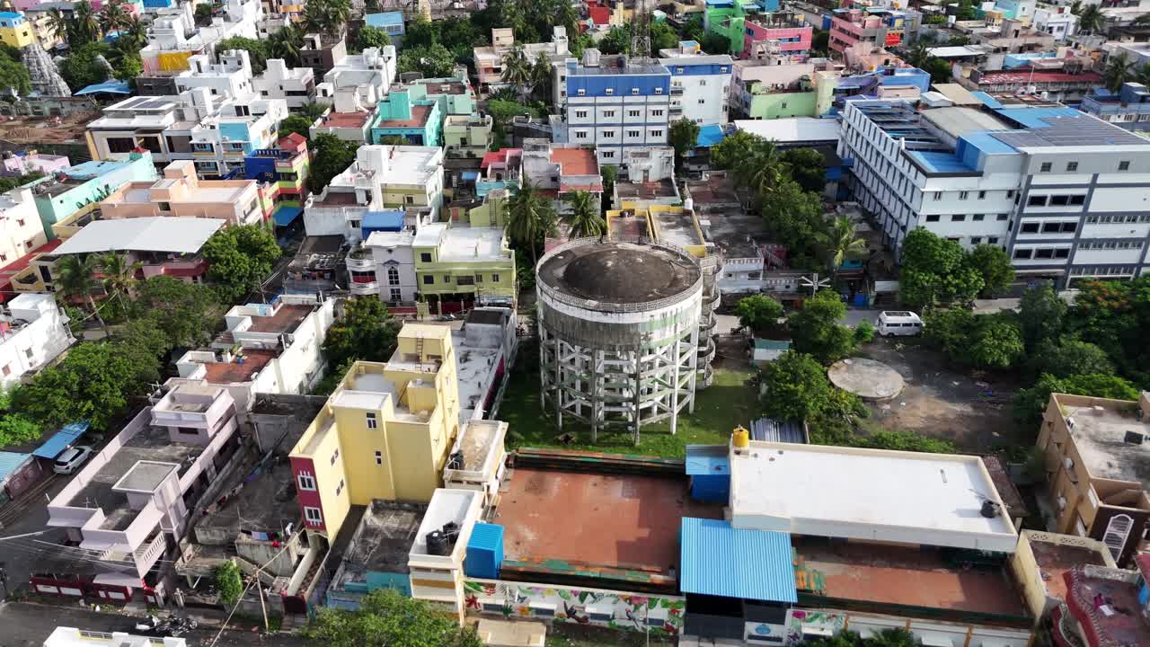 A high-angle aerial shot of a large, circular water tank in the center of a bustling residential neighborhood in India. The drone footage captures colorful houses, rooftops, and green trees