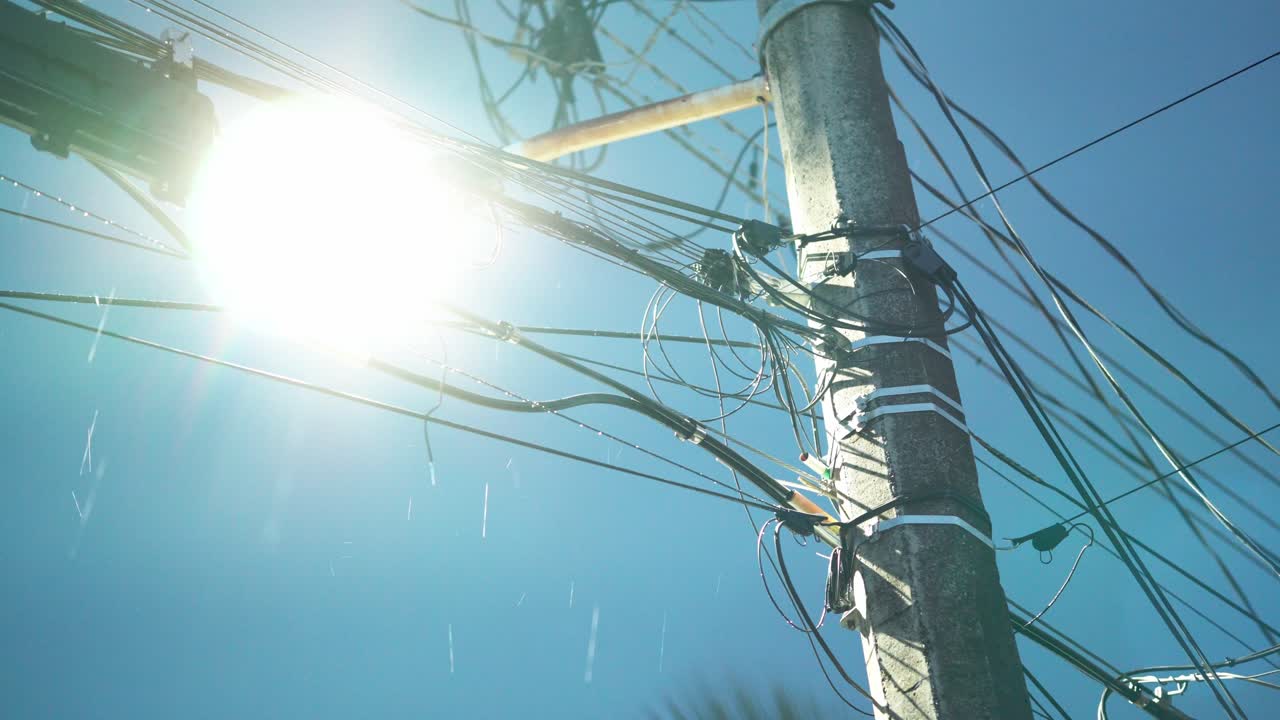 Close-up of an electric pole and tangled wires during rain, backlit by intense sunlight.