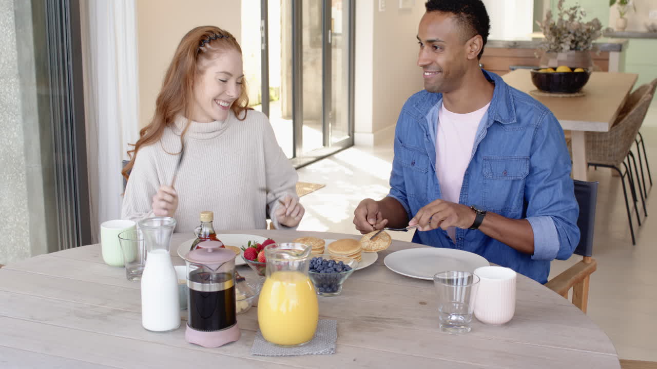 Diverse couple enjoying breakfast with pancakes and fruit, smiling at home