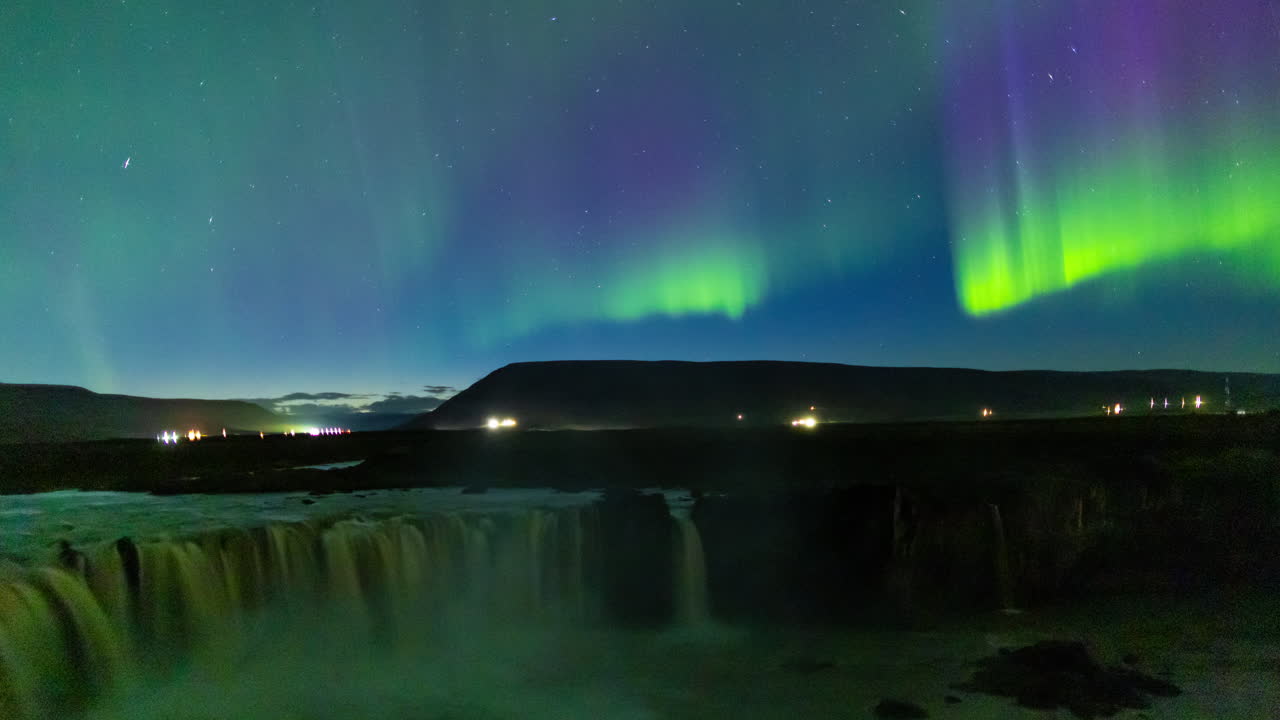 Aurora boreale alla cascata di Goðafoss, in Islanda. Spettacolari colonne verdi e rosse luminose nel cielo, con luci del traffico automobilistico e cascata in primo piano. Time lapse