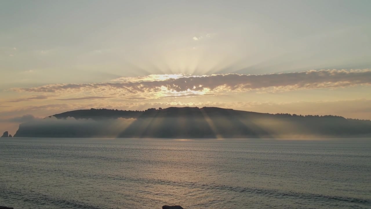 Revealing sun emerging from cloud, casting sun rays over island at rocky shoreline with mist