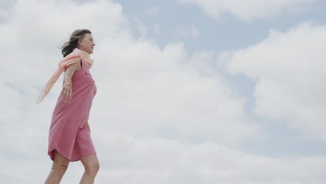 happy senior caucasian woman walking against cloudy sky on beach and raising arms, in slow motion