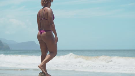 A-girl-in-a-bikini-stands-on-a-log-with-ocean-waves-and-mountains-in-the-background