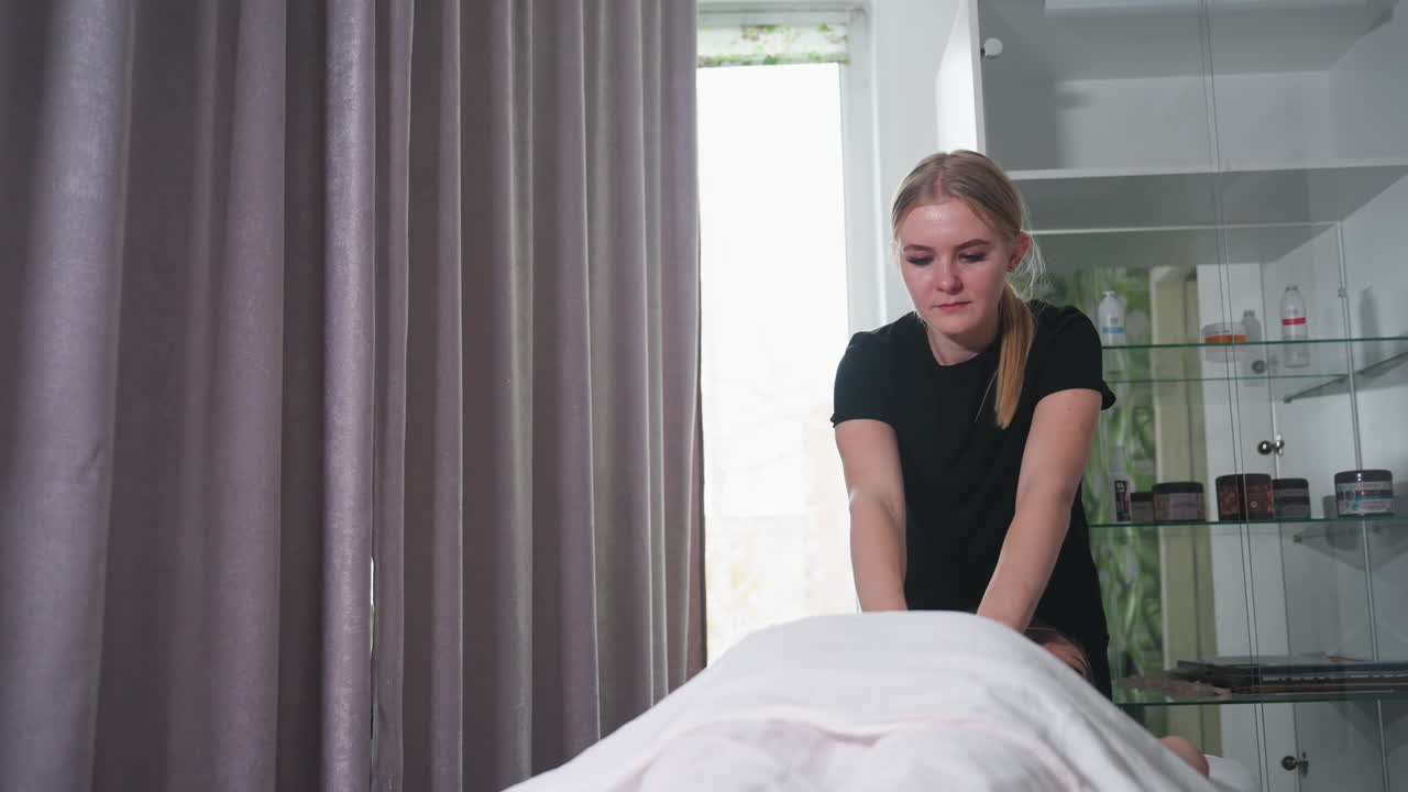 Massage therapist in black shirt performing treatment on client in bright wellness space, with gray curtains and clear cabinet shelves in background holding skincare and wellness products