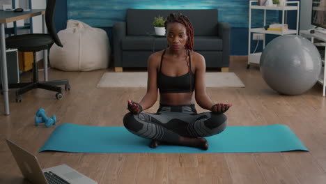 flexible slim black woman sitting in lotus position on yoga map in living room