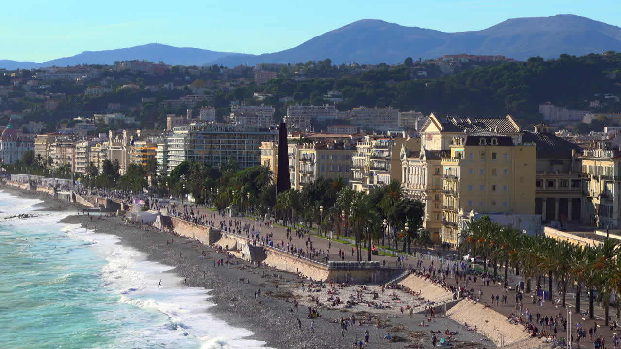 Turquoise waves crashing along the pebble beach of the Promenade des Anglais, with colorful buildings and palm trees lining the seafront