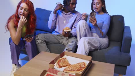amigos de la generación z sentados en el sofá comiendo pizza para llevar a casa y usando teléfonos móviles.
