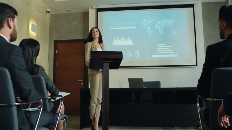Camera zoom in on caucasian businesswoman talking on a podium in a conference room and showing some charts and graphics on the big screen