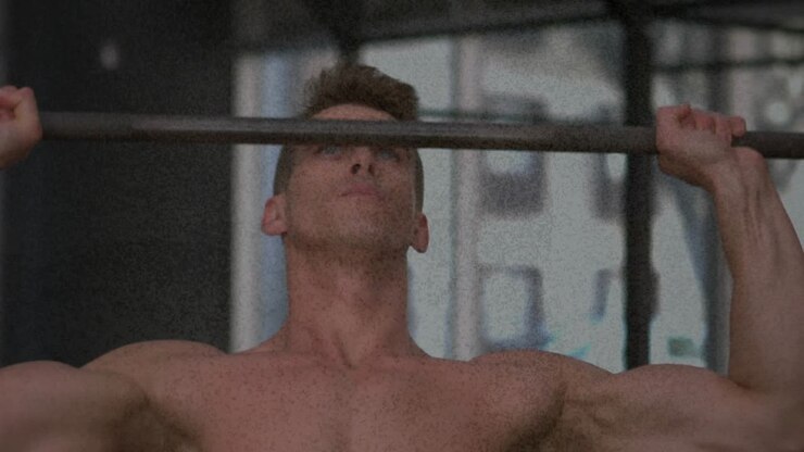 Shirtless male exerciser raising arms, gripping pull-up bar, pulling upward for strength training