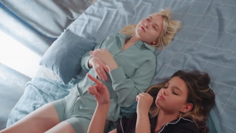 women lying on bed during relaxed indoor moment, with one in black pajamas singing expressively and using hand gestures while the other in green listens attentively