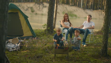 a family in nature parents watch as two boys at the fire roast marshmallows on sticks in the background of the tent. tent camp as a family