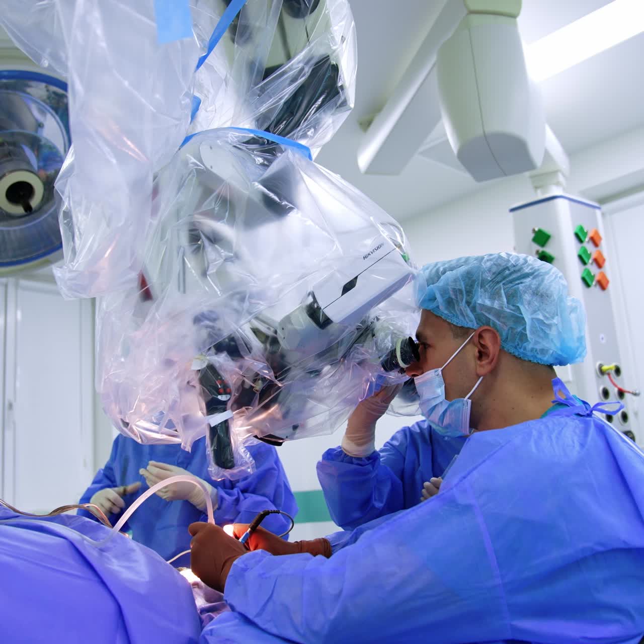 Male Caucasian neurosurgeon sitting at neuro microscope. Side view. Professional uses electric tools at procedure holding them in both hands