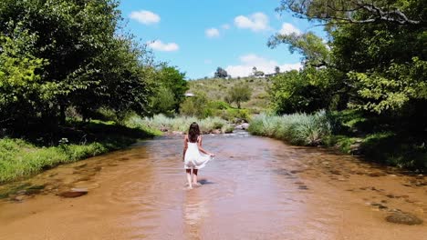 Wide-shot-of-a-woman-in-a-white-dress-walking-happily-through-a-fresh-water-stream-in-the-middle-of-the-forest,-normal-speed-version