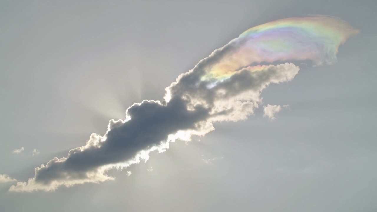 Long nuage dérivant montrant une frange irisée dans un ciel pâle, le soleil en contre-jour créant des rayons argentés.