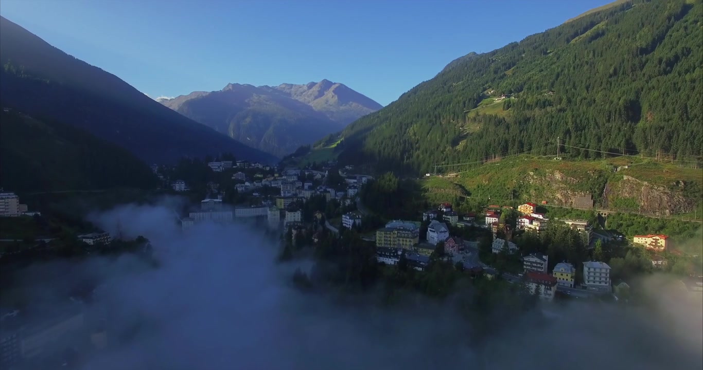 Lento movimento aereo rivela un compatto resort alpino austriaco con hotel, case, strade e pendii boscosi parzialmente nascosti da una fitta nebbia mattutina sotto aspre cime montuose
