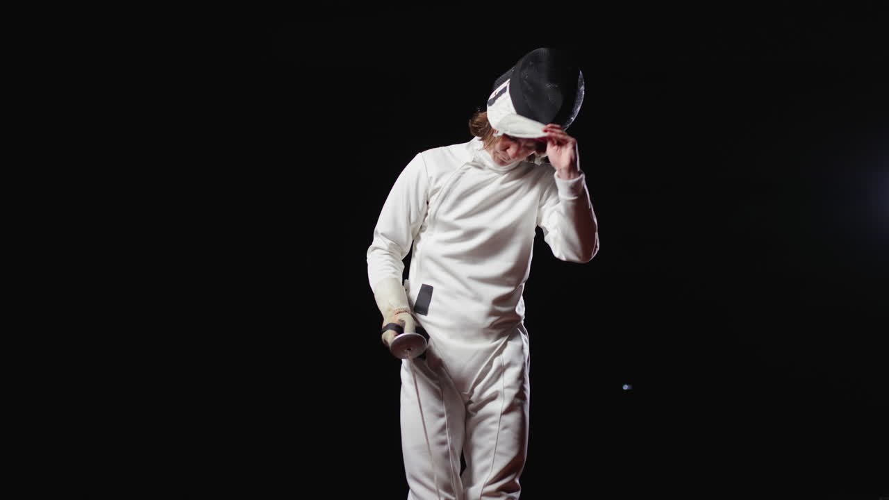 A male fencer in a white suit, holding a foil and wearing a mask, practices in a dark room.