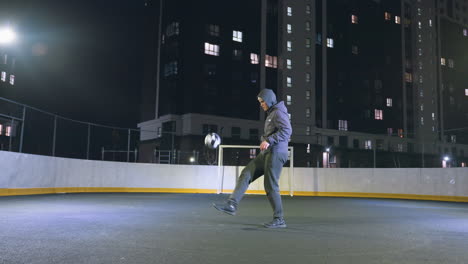 man joggling football under bright urban lights on outdoor sports field during nighttime, background features illuminated metallic fence, yellow and red vibrant lighting