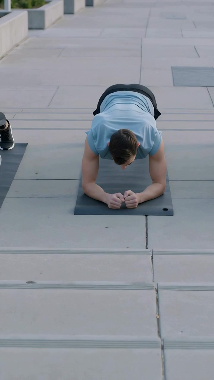 Vertical video: Doing elbow plank, man in blue shirt toning core on dark plaza mat