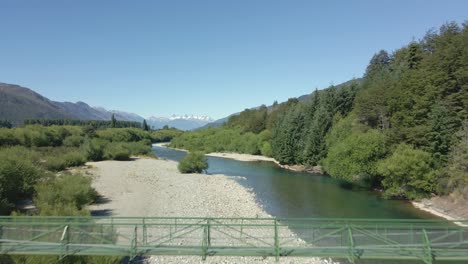 Aerial-dolly-in-of-river-and-elevated-bridge-surrounded-by-pine-trees-and-mountains,-Pasarela-Rio-Azul,-Patagonia-Argentina