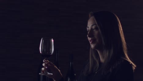 Mujer-Morena-Levantando-Una-Copa-De-Vino-Tinto-En-Un-Estudio-Romántico-De-Ambiente-Oscuro,-Mirando-Directamente-A-La-Cámara