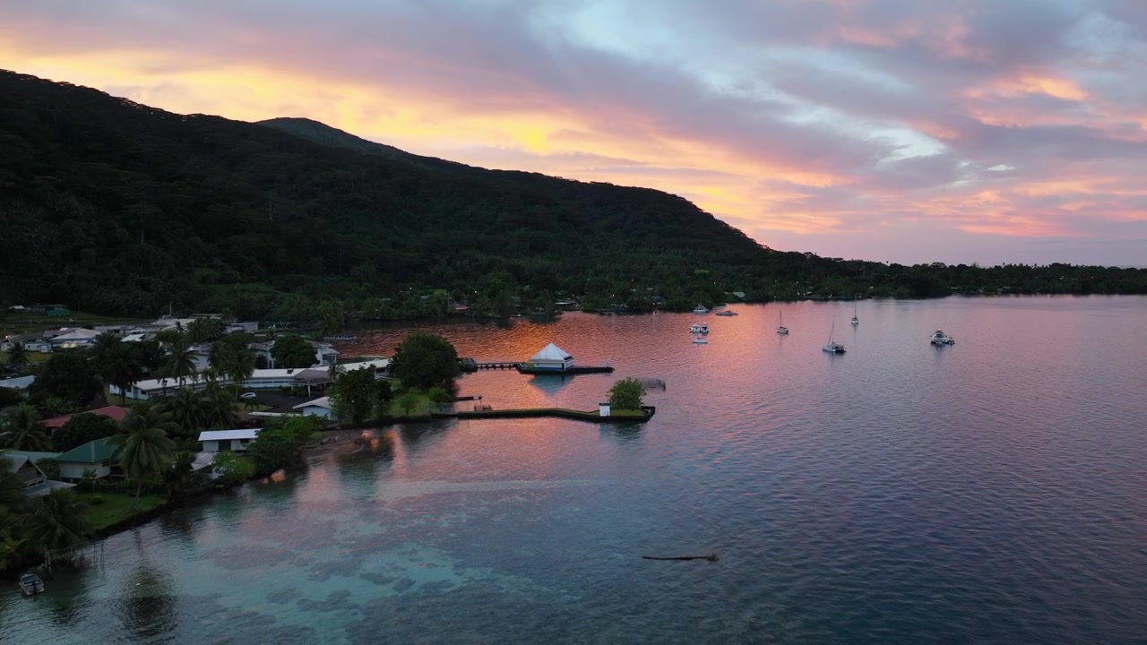 Piccole barche galleggiano su una laguna calma accanto a un villaggio costiero fiancheggiato da palme all'alba a Tahiti, Polinesia Francese; la silhouette di una montagna si erge sotto un cielo arancione brillante all'alba. Ampia ripresa aerea con drone.