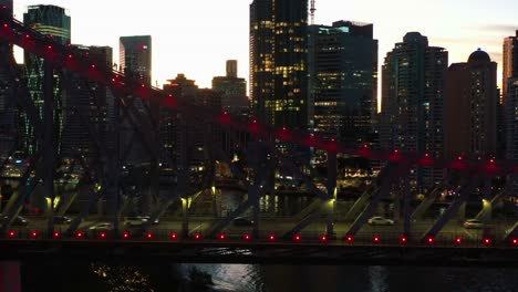 Escalada-De-Aventura-En-El-Puente-De-La-Historia-Icónica,-Gente-Subiendo-A-Un-Puente-Voladizo-Con-Mucho-Tráfico-Cruzando-El-Río-Al-Atardecer-Y-El-Paisaje-Urbano-Iluminado-Del-Centro-De-La-Ciudad-En-El-Fondo,-Toma-Aérea-De-La-Ciudad-De-Brisbane