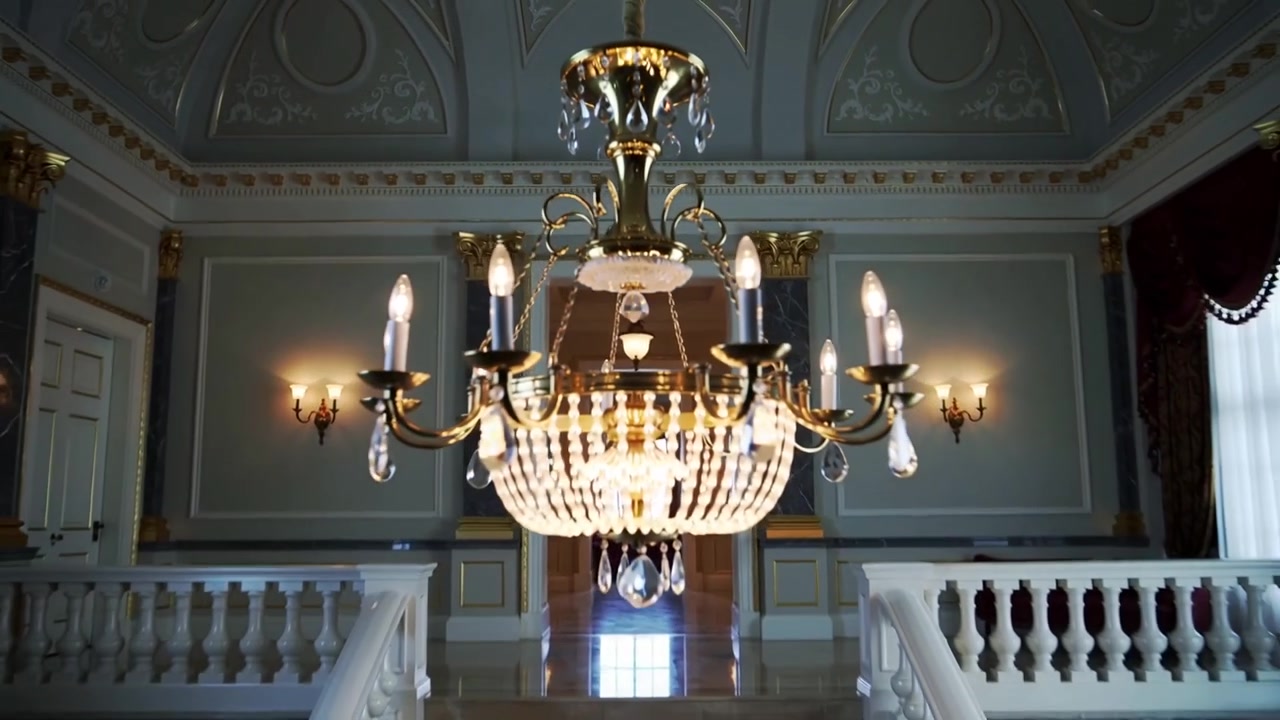 Telecamera che si allontana rivelando un lampadario di cristallo sopra una grande scala doppia, esponendo un soffitto ornato.