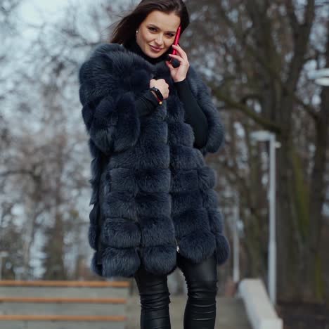 luxury woman in fur coat. beautiful young model walking in the park with a phone and looking at camera in cold season. slow motion.