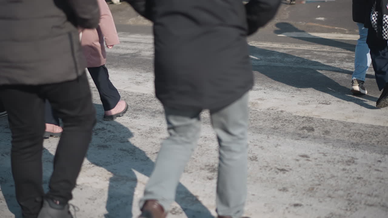 low angle leg view of people in winter jackets with backpacks crossing city street, boots stepping, long shadows on sunny asphalt, side movement during morning commute, crowd flow, urban pedestrians