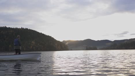 Junger-Mann-Steht-Auf-Einem-Kleinen-Boot,-Das-Bei-Sonnenuntergang-Ein-Fischernetz-Auf-Dem-See-Wirft