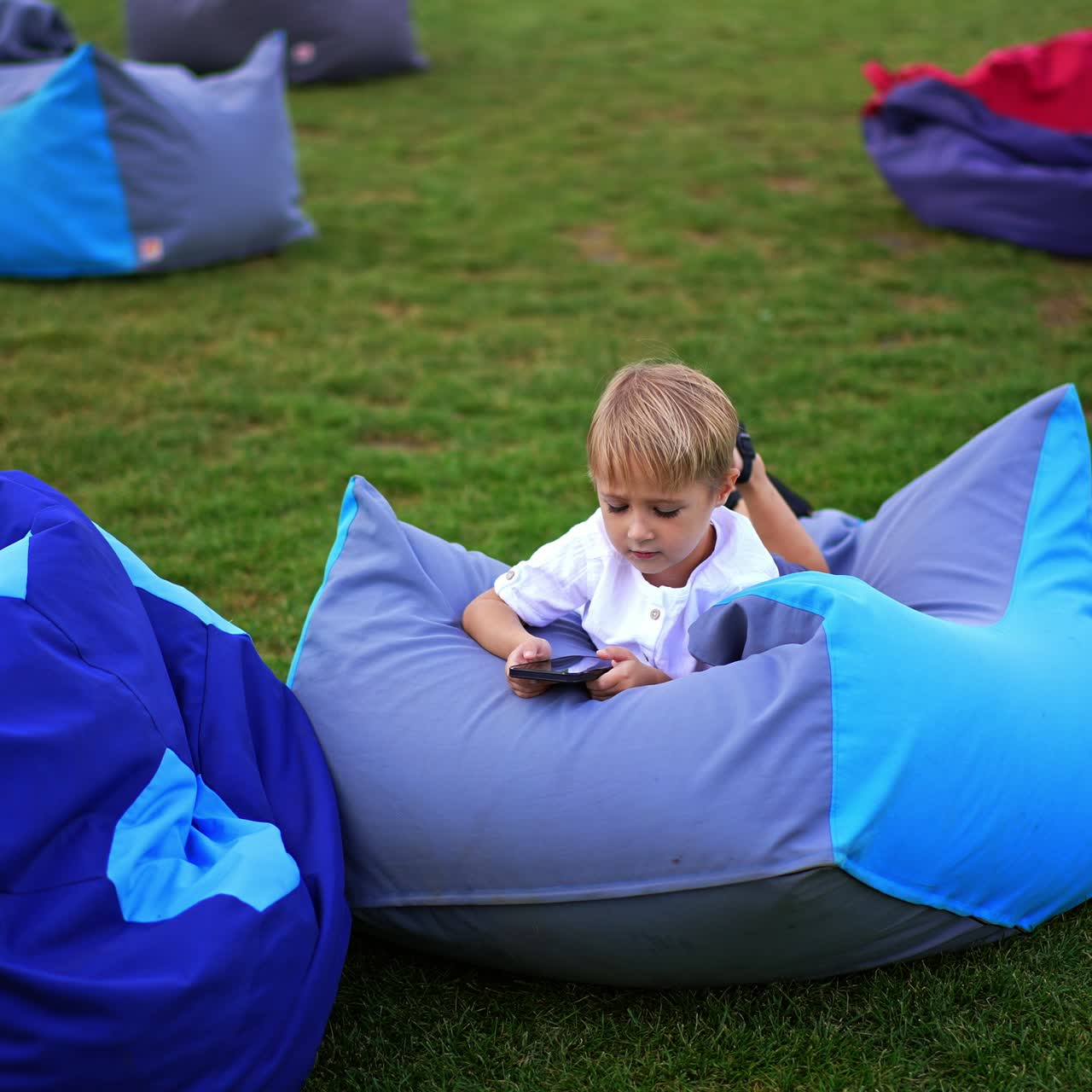 Green lawn with few bean bag chairs. Little blond boy lies on one of the chairs holding a smartphone in hands