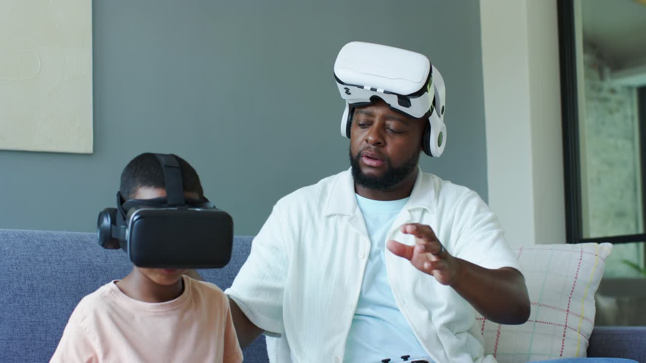 Afroamerikansk far og søn i sofaen; sønnen sætter et VR-headset på, og faren justerer og guider med en controller.