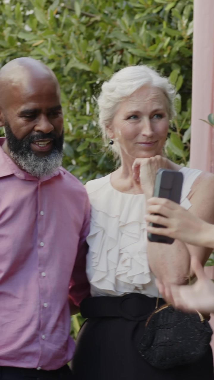 Video verticale: Ospiti di diverse etnie, uomo in rosa che si tiene a braccetto in giardino, telefono che riprende la sposa che entra.