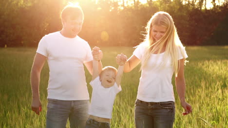 Madre,-Padre-E-Hijo-Caminan-Por-El-Campo-Con-Púas-En-Camisetas-Blancas-Y-Jeans-Divertidos-Balanceándolos-En-Sus-Manos