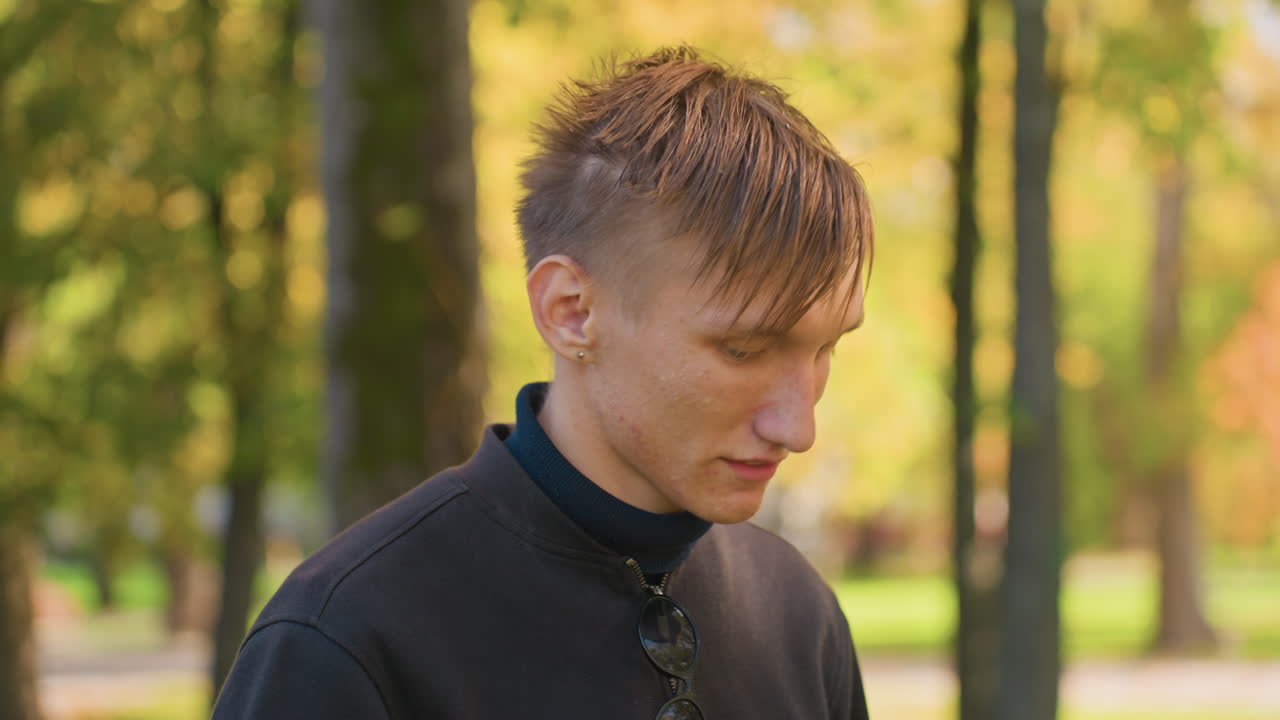 young man drinking water in park during autumn damp hair after run, dark sweater, clear plastic bottle visible, long sip showing relief, golden leaves, treelined path, candid closeup, calming
