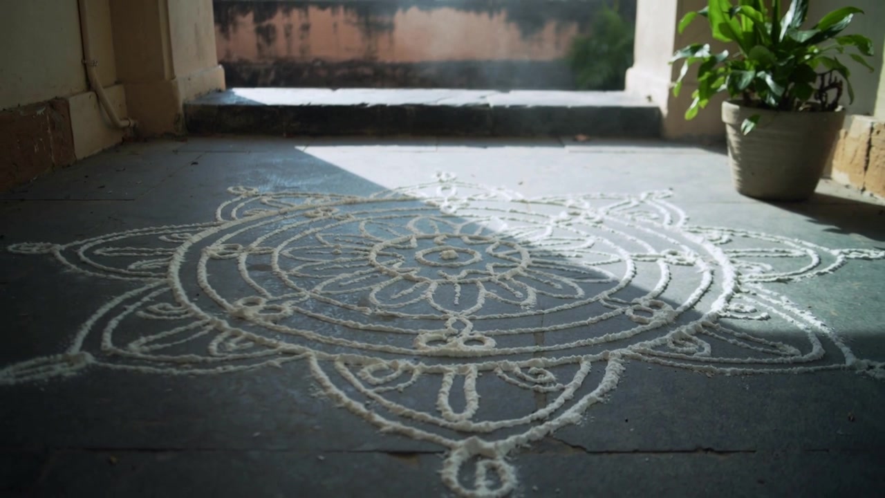 Un raggio di sole si sposta sul portico al cambiare dell'angolazione del sole, illuminando un rangoli sul pavimento di cemento e una pianta.