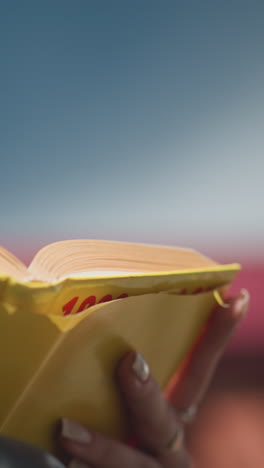 primer plano de una mujer ligeramente borrosa hojeando páginas de un libro de portada amarilla en su mano, con enfoque en el borde del libro y colores vibrantes, fondo de luz al aire libre brillante