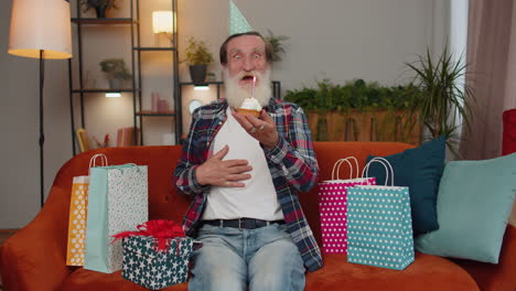 happy senior old man celebrating birthday party, makes wish blowing burning candle on small cupcake