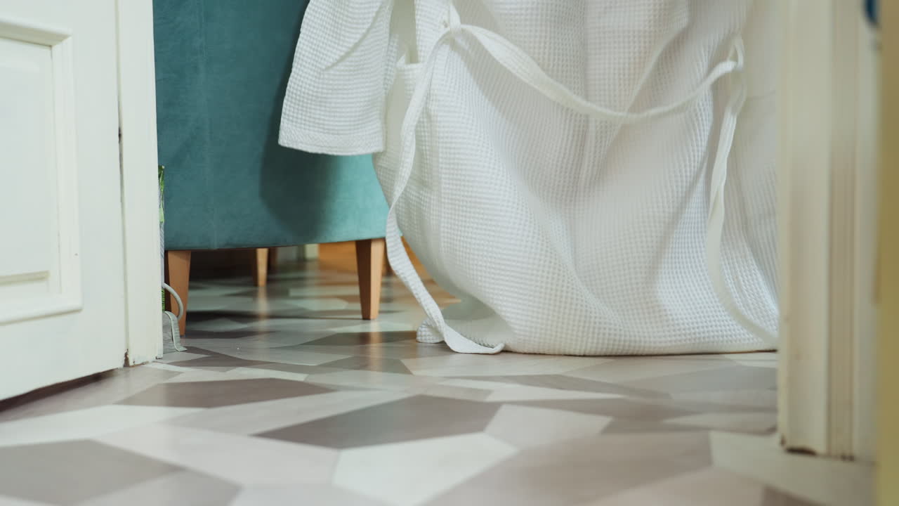 Light skin woman wearing white robe and soft white slippers standing near blue chair on geometric patterned floor, gently pulling robe, captured from low angle in calm indoor spa setting