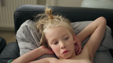 in a close-up shot, a 7-year-old caucasian boy with long hair in a bun lies on a black couch, covered in a blanket