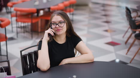 joven con gafas involucrada en una conversación telefónica, mirando hacia la derecha, sentada en el patio de comidas del centro comercial con un fondo borroso de estructuras modernas, mesas y sillas