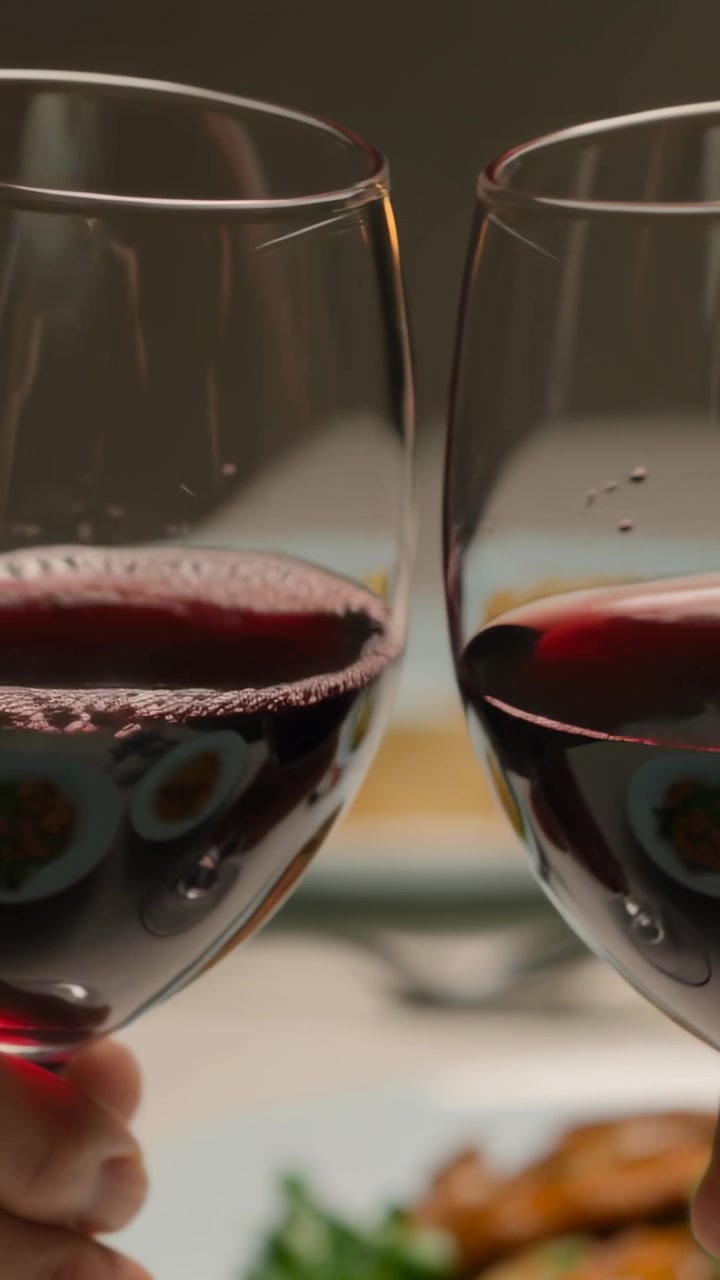 Video verticale: Due calici di vino rosso che si scontrano mentre le mani brindano durante la cena, con i piatti sfocati.