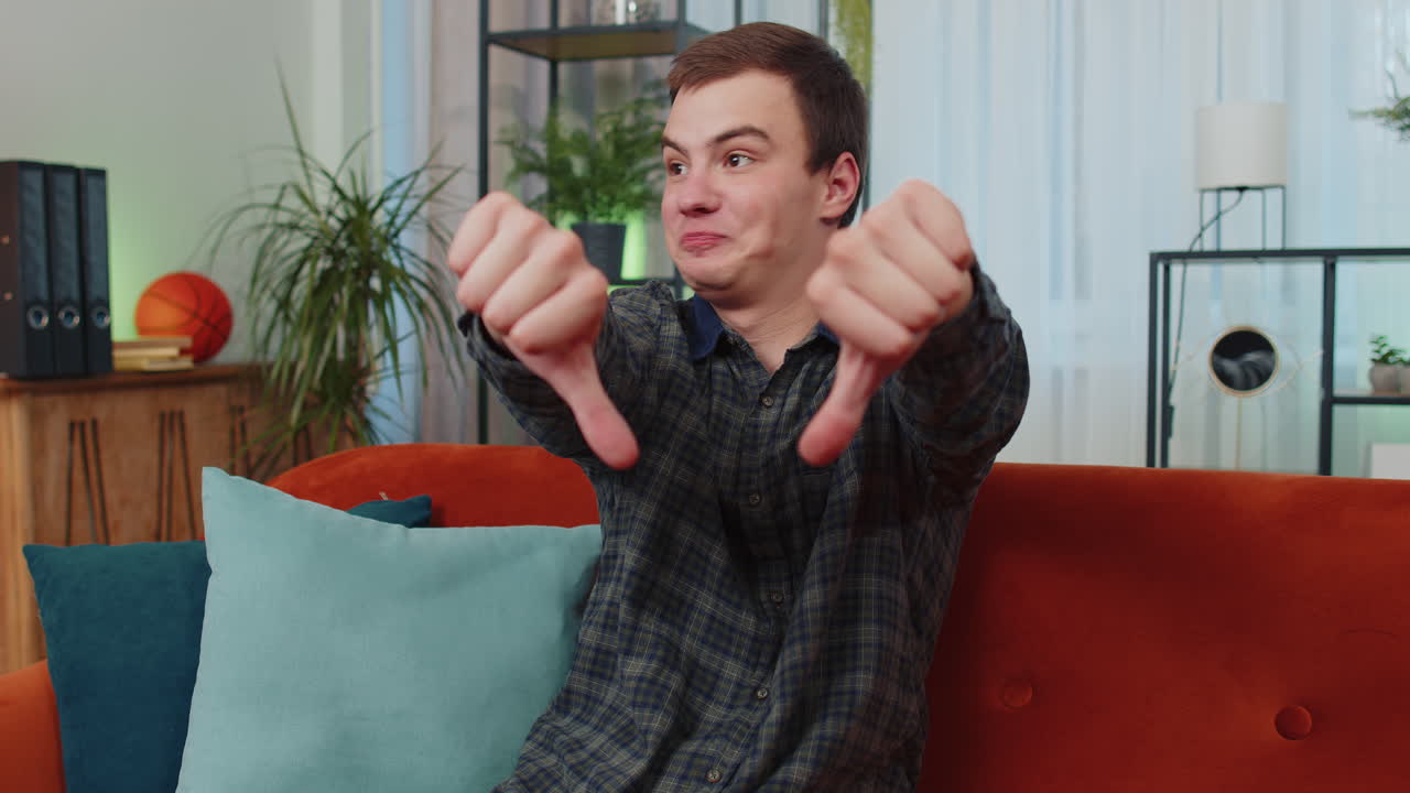 화가 난 젊은 남자가 엄지손가락을 내려놓고, 나쁜 일을 싫어하고, 승인하지 않고, 집에서 불만족스러운 피드백을 합니다.