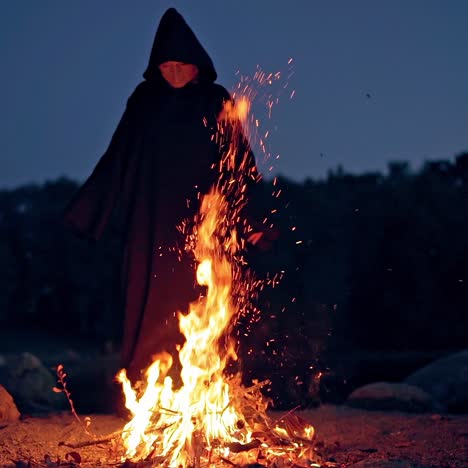 witch makes scary ritual in the fire. mysterious wizard in black cloak doing shamanism over the bright fire flame in nature at night.