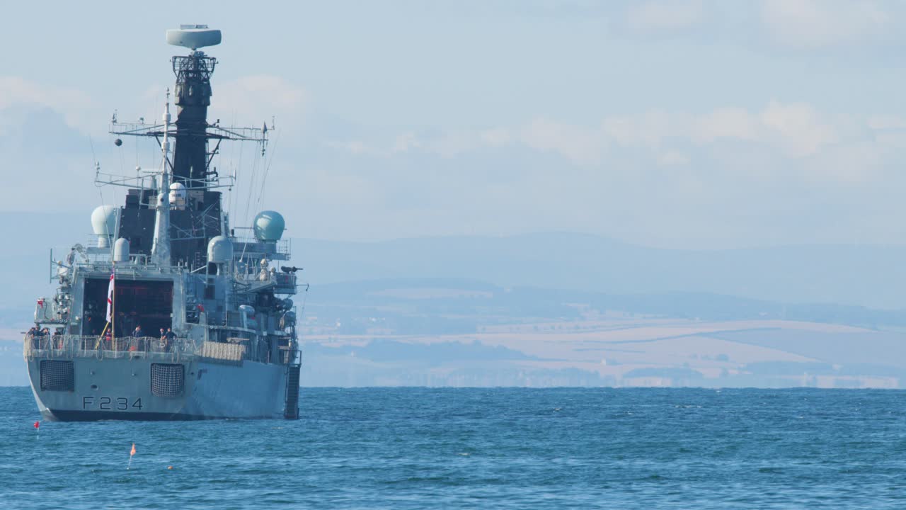 A large military vessel steadily advances toward the camera across tranquil blue waters, with distant coastline and hills visible under bright daylight