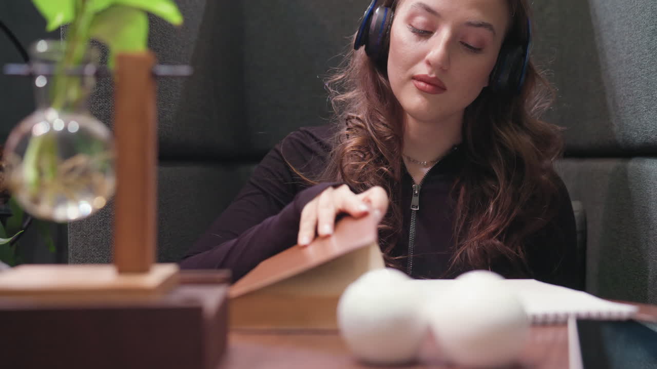 Lady with curly brown hair wears black headset with eyes closed, enjoying calm moment while seated near closed brown book and notebook on wooden table with soft lighting and minimal decoration