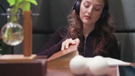 lady with curly brown hair wears black headset with eyes closed, enjoying calm moment while seated near closed brown book and notebook on wooden table with soft lighting and minimal decoration