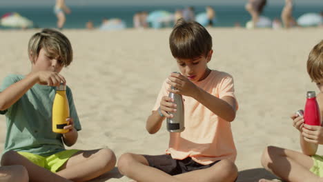 slider shot of boys sitting on sandy beach and drinking water