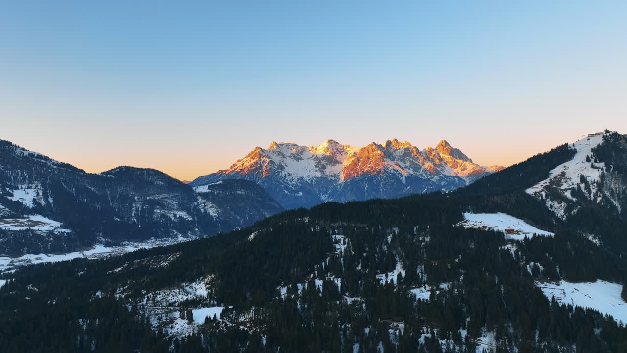 The drone is flying over a mountain with trees towards the big mountain lit by the golden sun during sunset in Austria Aerial Footage 4K
