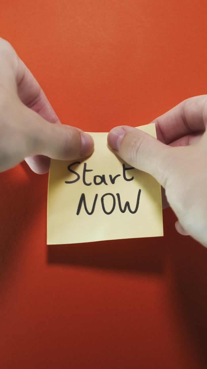 Hands posting Start Now sticky note on orange wall, evoking urgency